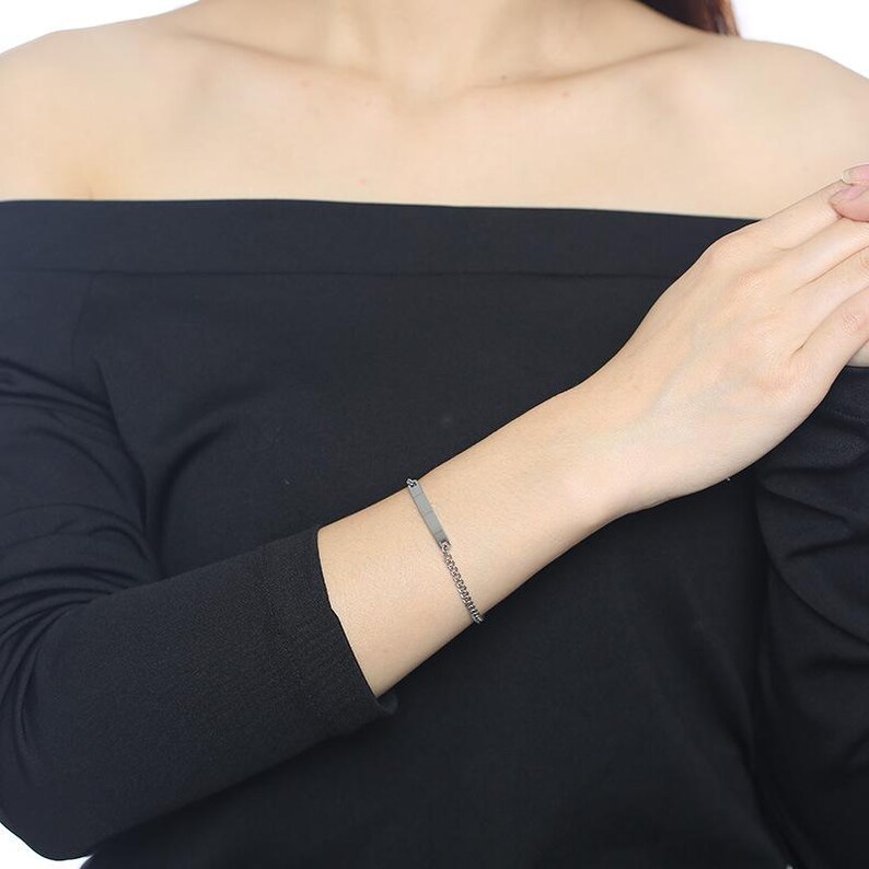 May include: A silver-toned metal chain bracelet featuring a small, rectangular identification plate. The bracelet consists of a delicate link chain that connects to the flat, smooth metal bar, creating a simple and clean design for the wrist.
