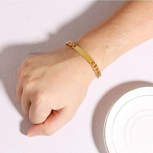 May include: A gold-toned chain link bracelet featuring a smooth, rectangular identification plate in the center. The bracelet is constructed with interlocking metal links that connect to the flat, polished bar, creating a continuous loop design.