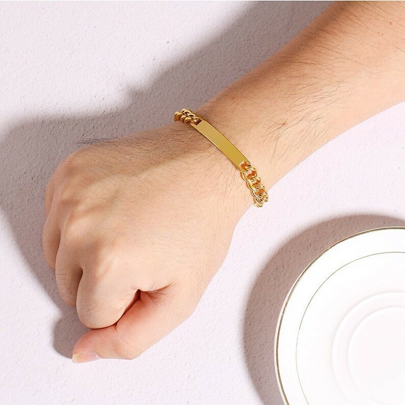 May include: A gold-toned chain link bracelet featuring a smooth, rectangular identification plate in the center. The bracelet is constructed with interlocking metal links that connect to the flat, polished bar, creating a continuous loop design.