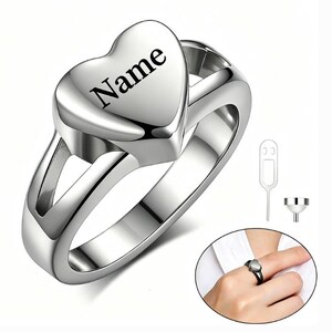 May include: A silver-toned metal ring featuring a heart-shaped top with the word Name engraved in black. The ring has a split shank design and is accompanied by a small metal funnel and a thin metal pin tool for filling the hollow heart compartment.