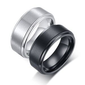 Puede incluir: Dos anillos, uno plateado y otro negro, se muestran sobre un fondo blanco. Los anillos tienen una superficie metálica plana y cepillada con un borde pulido. El anillo negro tiene un interior brillante.