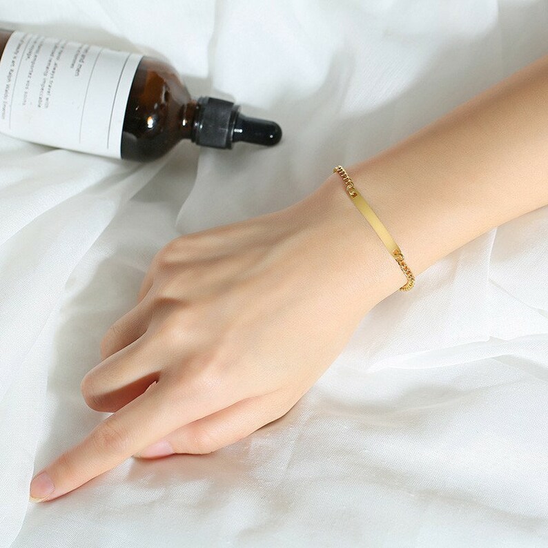 May include: A gold-toned bracelet featuring a thin, flat, rectangular metal bar centerpiece connected to a delicate chain. The bracelet is worn on a wrist, showcasing the polished metallic finish and the simple, streamlined design of the jewelry piece.