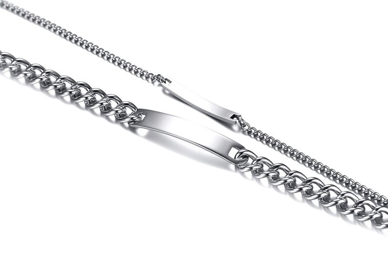 May include: Two silver-toned metal bracelets featuring a polished, curved identification plate at the center. Each bracelet is constructed with a classic curb chain link design, showcasing a reflective, smooth surface and a metallic finish.