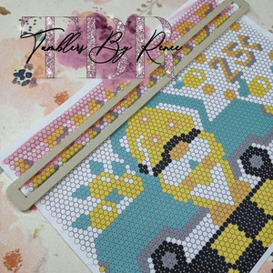 May include: A close-up of a colorful beaded design of two construction vehicles on a white background. The design is partially covered by a white plastic tool used for beading.