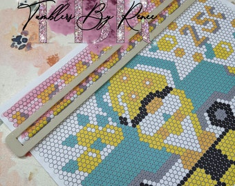 May include: A close-up of a colourful beaded design of two construction vehicles on a white background. The design is partially covered by a white plastic tool used for beading.