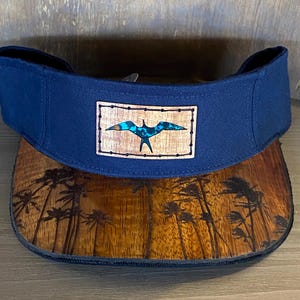 May include: A navy blue baseball cap with a wood panel brim featuring a palm tree design. The front of the cap has a wood patch with a bird design.