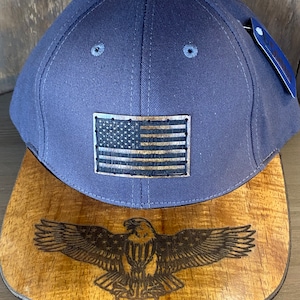 May include: A navy blue baseball cap with a wood-look American flag patch on the front. The cap has a wood brim with a laser-etched bald eagle design.
