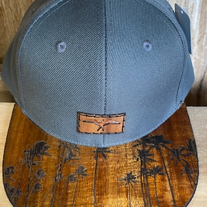 May include: A gray and brown baseball cap with a wood-look brim featuring a palm tree design. The front of the cap has a brown leather patch with a bird design and the text "Albatross".