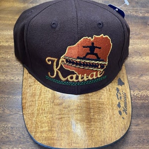 Op de afbeelding: Bruine baseballpet met een rand die eruitziet als hout. De voorkant van de pet heeft een geborduurd ontwerp van het eiland Kauai met een persoon die een yogapose doet en het woord "Kauai" in een gestileerd lettertype. De rand heeft het woord "Hawaii" erop geschreven.