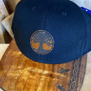 May include: A black baseball cap with a brown leather tree of life patch on the front. The bill of the cap is made of wood with a carved design of two fish and a Celtic knot.