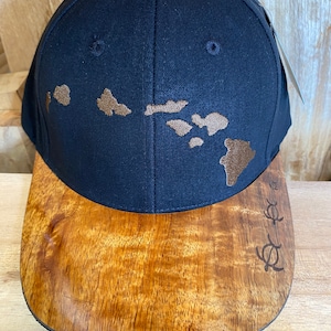 May include: A black baseball cap with a brown embroidered outline of the Hawaiian islands. The brim of the cap is made of wood with a brown wood grain pattern and a carved turtle design.
