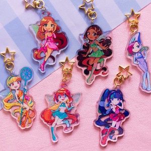 Puede incluir: Conjunto de siete amuletos de personajes de dibujos animados coloridos con llaveros de estrella dorada. Cada amuleto presenta un personaje de hada diferente con alas y un diseño único.