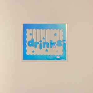 May include: A blue card with a white scalloped border and the word "drinks" in white letters with a blue outline. There are small white stars scattered around the card.