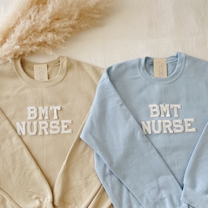 May include: Two crewneck sweatshirts, one tan and one light blue, are displayed. Each sweatshirt has the text "BMT NURSE" in white, block-style lettering. The sweatshirts are made of a soft material and are likely for medical professionals.
