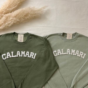 Puede incluir: Dos sudaderas de manga larga con la palabra "CALAMARI" en letras blancas en la parte delantera. La sudadera de la izquierda es verde militar y la sudadera de la derecha es verde salvia.