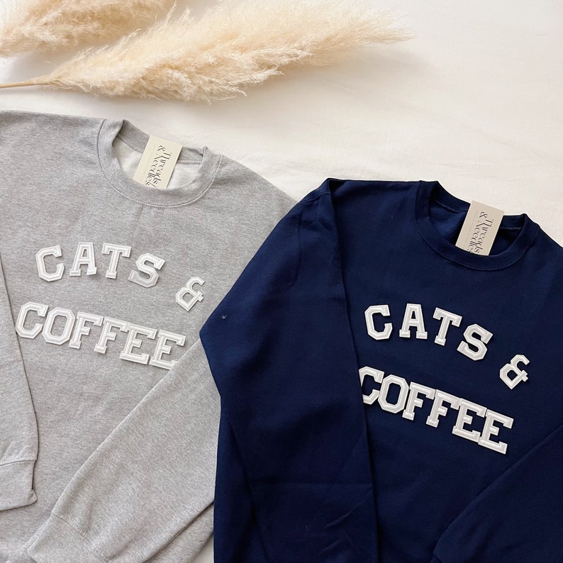 May include: Two long-sleeved sweatshirts with the text "Cats & Coffee" in white embroidered letters. One sweatshirt is gray and the other is navy blue.