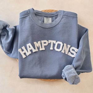 Op de afbeelding: Een blauw sweatshirt met een wit "HAMPTONS" logo in een lettertype in collegiale stijl.