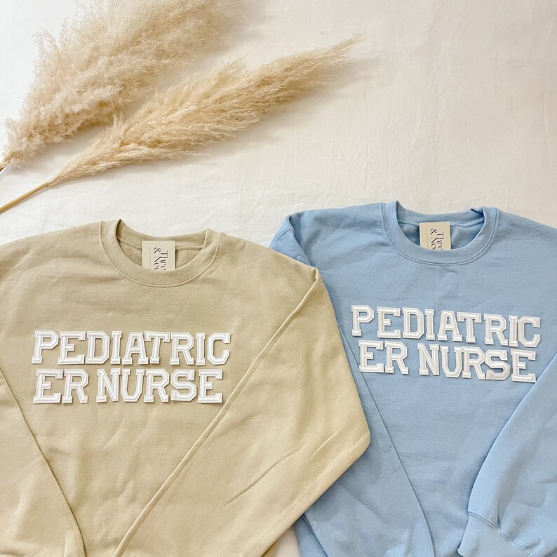 May include: Two long-sleeved sweatshirts with the text "Pediatric ER Nurse" in white letters. One sweatshirt is beige and the other is light blue.