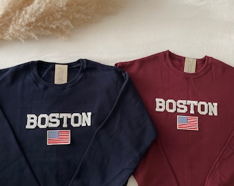May include: Two long-sleeved sweatshirts with the word "BOSTON" in white letters and an American flag patch. The sweatshirt on the left is navy blue and the sweatshirt on the right is maroon.