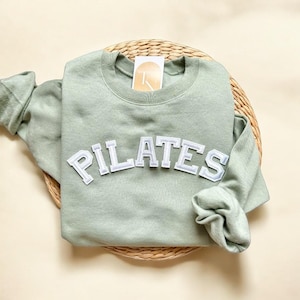 May include: Sage green sweatshirt with white "PILATES" lettering in a varsity style font.