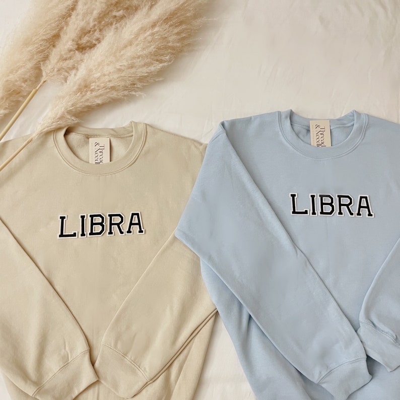 May include: Two sweatshirts with the word "LIBRA" in black block letters on the front. One sweatshirt is beige and the other is light blue.