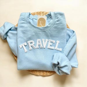 Peut inclure: Un sweat-shirt bleu clair avec le mot "TRAVEL" en lettres blanches sur le devant.