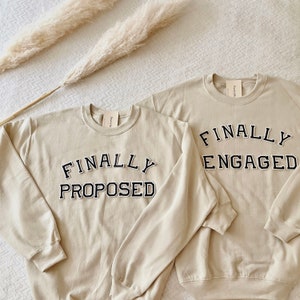 Peut inclure: Deux sweat-shirts beiges à col rond, l'un avec l'inscription "FINALLY PROPOSED" et l'autre "FINALLY ENGAGED" en lettres noires de style universitaire. Les sweat-shirts ont un design simple, idéal pour les couples. Le fond comprend des plantes séchées.