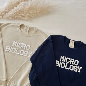 May include: Two crewneck sweatshirts, one beige and one navy blue, with the word "MICROBIOLOGY" in white, collegiate-style lettering. The sweatshirts have a tag that reads "Threads & Needles".
