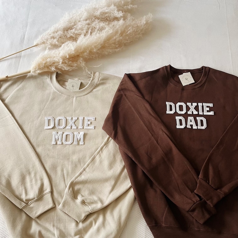 May include: Two long-sleeved sweatshirts, one beige and one brown, with white embroidered text. The beige sweatshirt says "DOXIE MOM" and the brown sweatshirt says "DOXIE DAD".