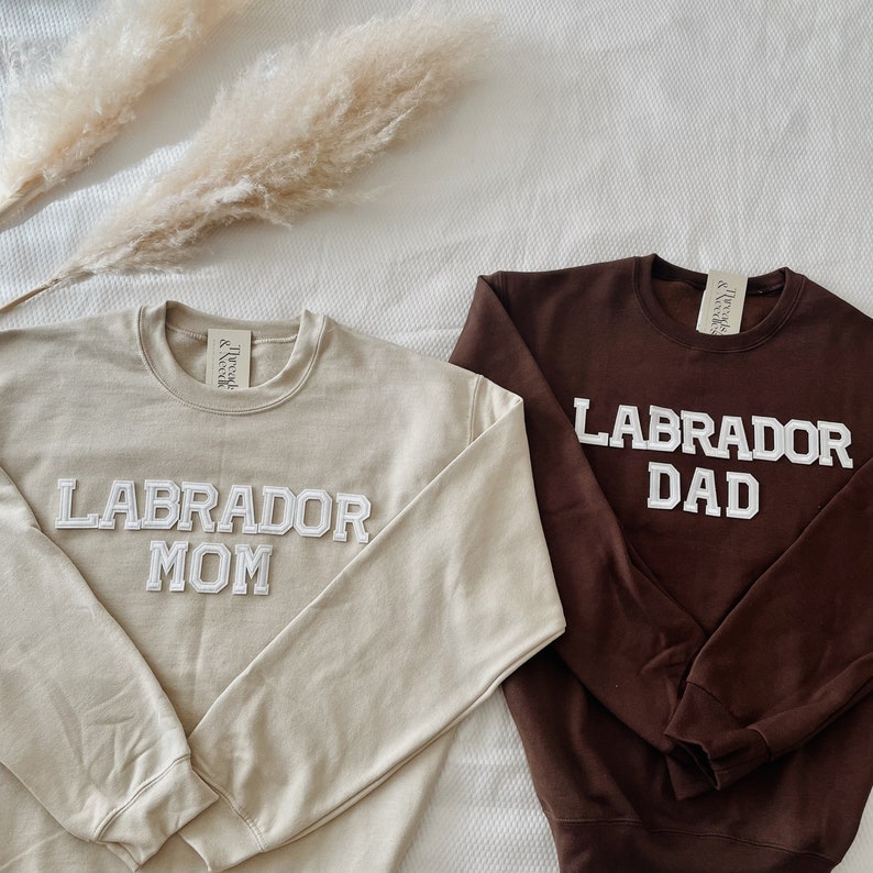 May include: Two crewneck sweatshirts. One is tan with "LABRADOR MOM" in white block letters. The other is brown with "LABRADOR DAD" in white block letters. Both have a tag that says "Thread & Nicole".
