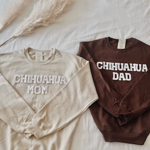 May include: Two crewneck sweatshirts, one tan and one brown, with the words "CHIHUAHA MOM" and "CHIHUAHUA DAD" in white, block lettering. The sweatshirts are displayed on a white surface with dried ornamental grass.