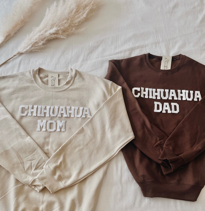 May include: Two crewneck sweatshirts, one tan and one brown, with the words "CHIHUAHA MOM" and "CHIHUAHUA DAD" in white, block lettering. The sweatshirts are displayed on a white surface with dried ornamental grass.