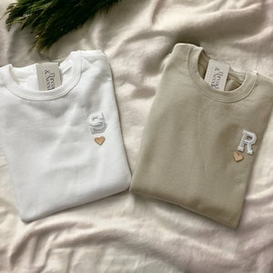 Puede incluir: Dos camisetas blancas y beige dobladas con el texto "Thread & Need" y un coraz&oacute;n y una letra bordada en cada una.