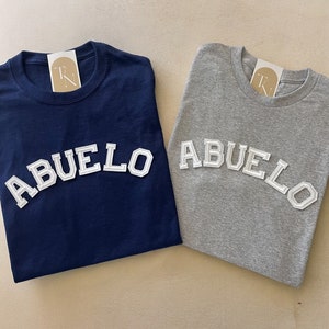 May include: Two t-shirts with the word "ABUELO" in white letters on a navy blue and a gray shirt.