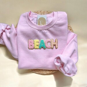May include: A pale pink sweatshirt featuring the word "BEACH" in colourful, textured embroidery. The garment is folded on a woven basket. A small tag is visible near the neckline of the sweatshirt.