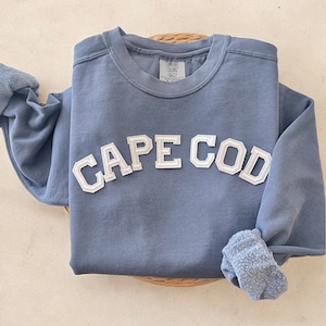 Op de afbeelding: Een blauw sweatshirt met witte geborduurde letters die "CAPE COD" lezen.
