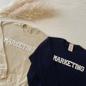 Op de afbeelding: Twee longsleeve sweatshirts met het woord "MARKETING" in witte reli&euml;fletters. E&eacute;n sweatshirt is beige en de andere is donkerblauw.