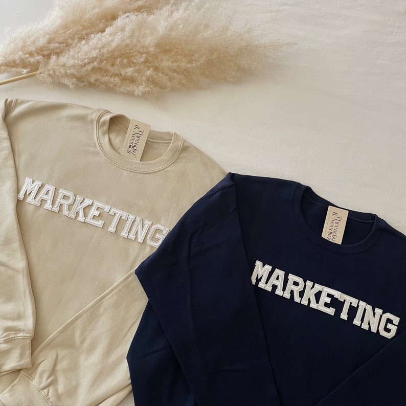 Op de afbeelding: Twee longsleeve sweatshirts met het woord "MARKETING" in witte reli&euml;fletters. E&eacute;n sweatshirt is beige en de andere is donkerblauw.