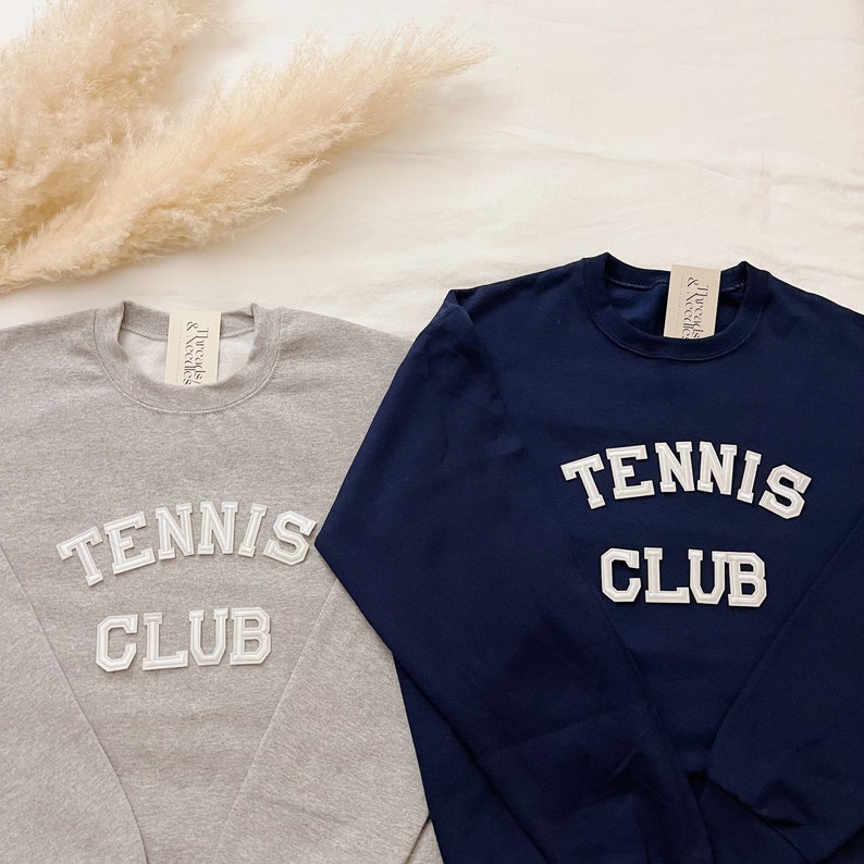 May include: Two long-sleeved sweatshirts with the text "Tennis Club" in white letters. One sweatshirt is grey and the other is navy blue.