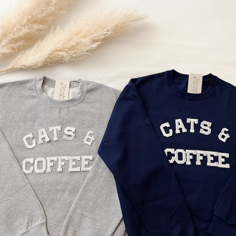 May include: Two sweatshirts, one gray and one navy blue, with the text "CATS & COFFEE" in white block letters.