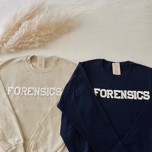 May include: Two sweatshirts with the word "FORENSICS" in white embroidered letters. One sweatshirt is beige and the other is navy blue.