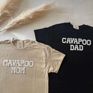 Op de afbeelding: Twee shirts met korte mouwen, één beige en één zwart, met de woorden "CAVAPOO MOM" en "CAVAPOO DAD" in witte blokletters. Elk shirt heeft een label met "Threads & Needle".