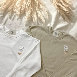 Puede incluir: Dos camisas de manga larga con letras y corazones bordados. La camisa de la izquierda es blanca con una "S" dorada y un peque&ntilde;o coraz&oacute;n rosa. La camisa de la derecha es marr&oacute;n claro con una "R" dorada y un peque&ntilde;o coraz&oacute;n rosa. Ambas camisas tienen una etiqueta blanca con el texto "& True & Co."