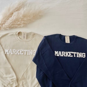 Op de afbeelding: Twee longsleeve sweatshirts met het woord "MARKETING" in witte reli&euml;fletters. E&eacute;n sweatshirt is beige en de andere is donkerblauw.