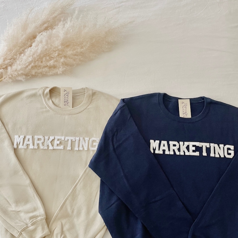 Op de afbeelding: Twee longsleeve sweatshirts met het woord "MARKETING" in witte reli&euml;fletters. E&eacute;n sweatshirt is beige en de andere is donkerblauw.