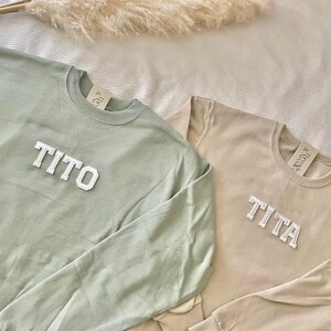 Puede incluir: Dos sudaderas de manga larga con los nombres "TITO" y "TITA" bordados en blanco en la parte delantera. La sudadera de la izquierda es verde salvia y la sudadera de la derecha es beige.