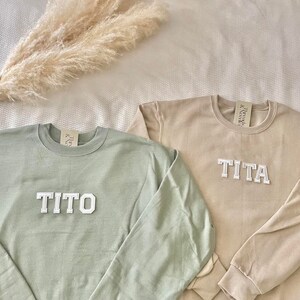 Puede incluir: Dos sudaderas de color claro con los nombres "TITO" y "TITA" en letras blancas en bloque. Las sudaderas est&aacute;n sobre una superficie blanca.