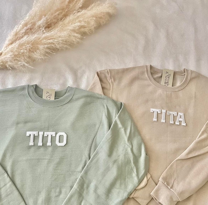 Puede incluir: Dos sudaderas de color claro con los nombres "TITO" y "TITA" en letras blancas en bloque. Las sudaderas est&aacute;n sobre una superficie blanca.