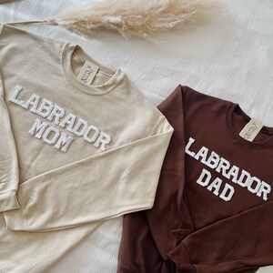May include: Two crewneck sweatshirts, one tan and one brown, with the words "LABRADOR MOM" and "LABRADOR DAD" embroidered in white. The sweatshirts have long sleeves and a tag on the upper back.