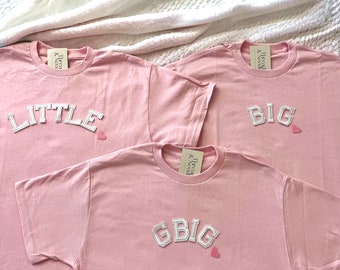 May include: Three pink t-shirts with white embroidered text. The shirts say "Little", "Big", and "GBig" with a small pink heart next to each word.